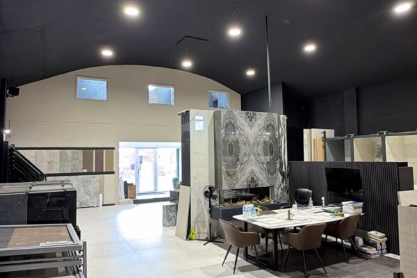 Interior showroom with marble slabs and sample materials, tables and chairs in foreground under a dark ceiling with bright lights on.