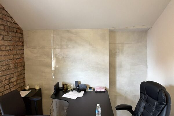 Office workspace with a black desk, two rolling chairs, papers and a water bottle on the desk; brick wall on the left and light tiled back wall.