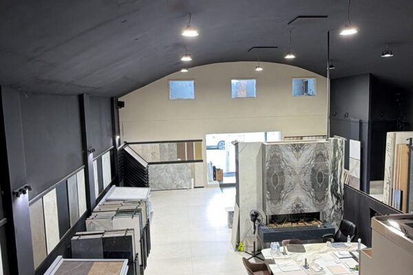 Tile and marble showroom interior with display racks along both walls, featuring sample slabs, and a central worktable under bright overhead lighting in a high curved ceiling space.