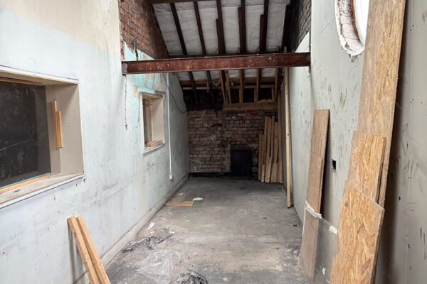 Narrow unfinished industrial space with exposed wooden roof beams, a skylight, and scattered plywood boards and debris along the floor.