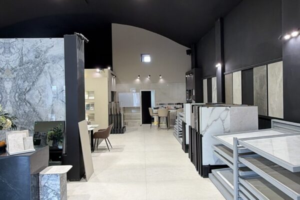 Interior of a tile showroom with white floor tiles, marble sample displays along the right, and a dark ceiling with recessed lights.