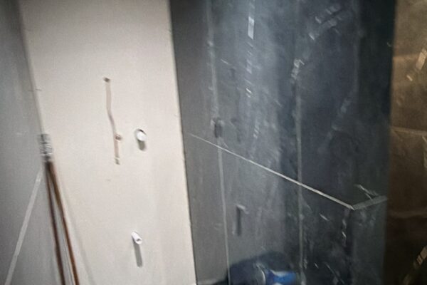Dark tiled bathroom under renovation with black marble walls, exposed pipes, and scattered debris on the floor.