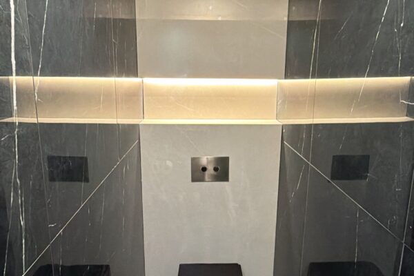 Modern black toilet set in a dark marble bathroom with recessed lighting and a wall-mounted flush panel above the seat area.